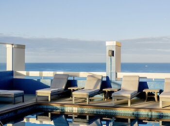 The image shows a coastal rooftop pool area with lounge chairs, a clear blue sky, and an ocean view in the background.