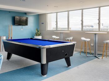 The image shows a modern recreation room with a pool table, bar stools, tables, a wall-mounted TV, and large windows letting in natural light.