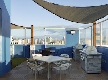 This image shows a rooftop patio with a table, chairs, and grills, overlooking a cityscape, shaded by fabric canopies on a sunny day.
