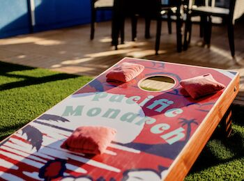 The image shows a colorful cornhole board with beanbags on it, set in an outdoor area with tables and chairs in the background.