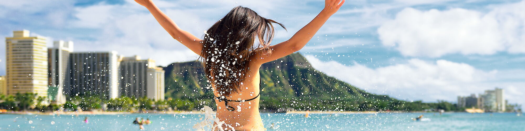 A person in a bikini splashing in the ocean, facing away from the camera with arms raised, city skyline and mountains in the background.