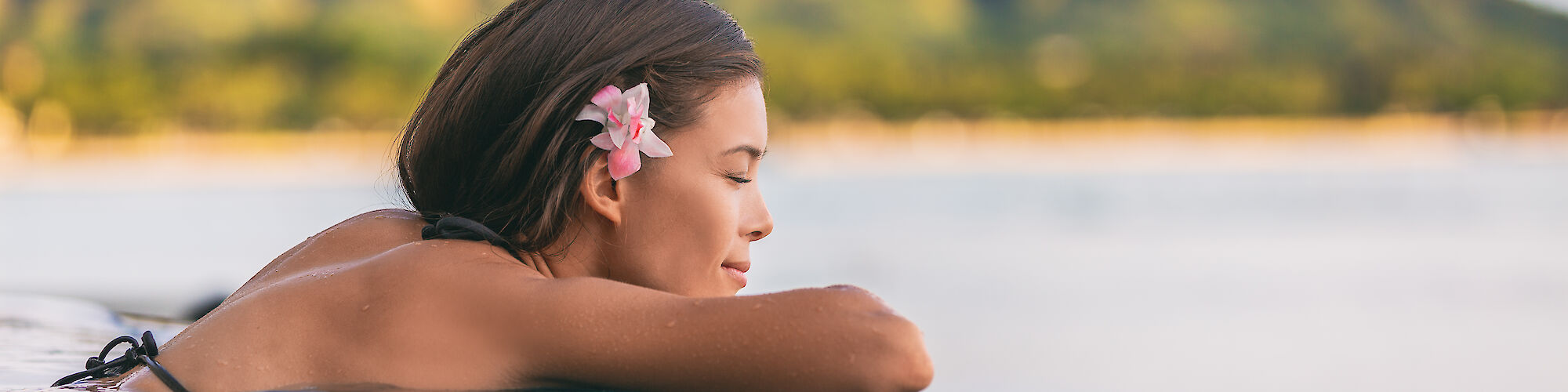 A woman with a flower in her hair is relaxing in a pool with a view of a mountain in the background at sunset.