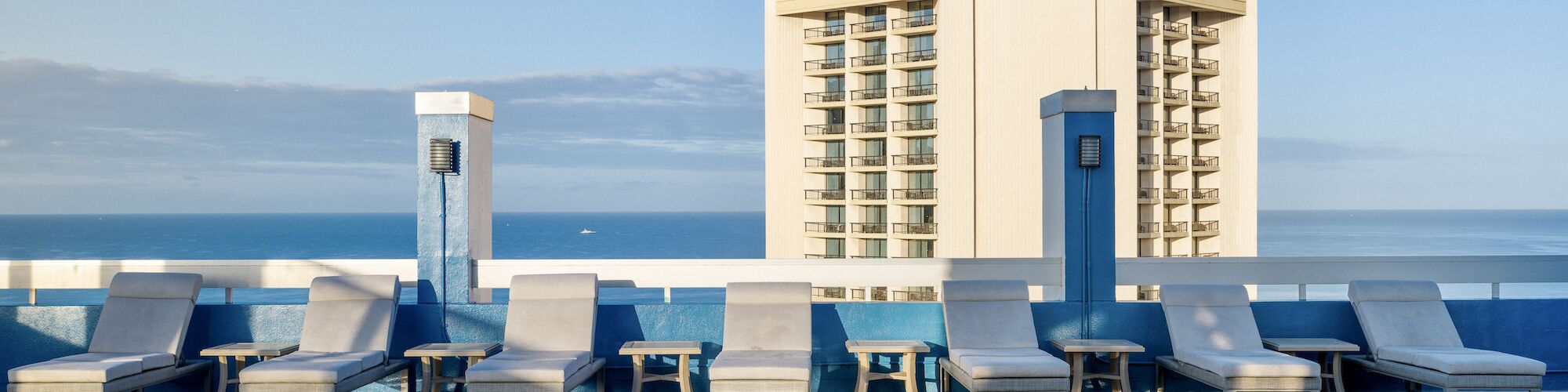 The image shows a rooftop pool area with lounge chairs, a body of water in the distance, and a tall building in the background.